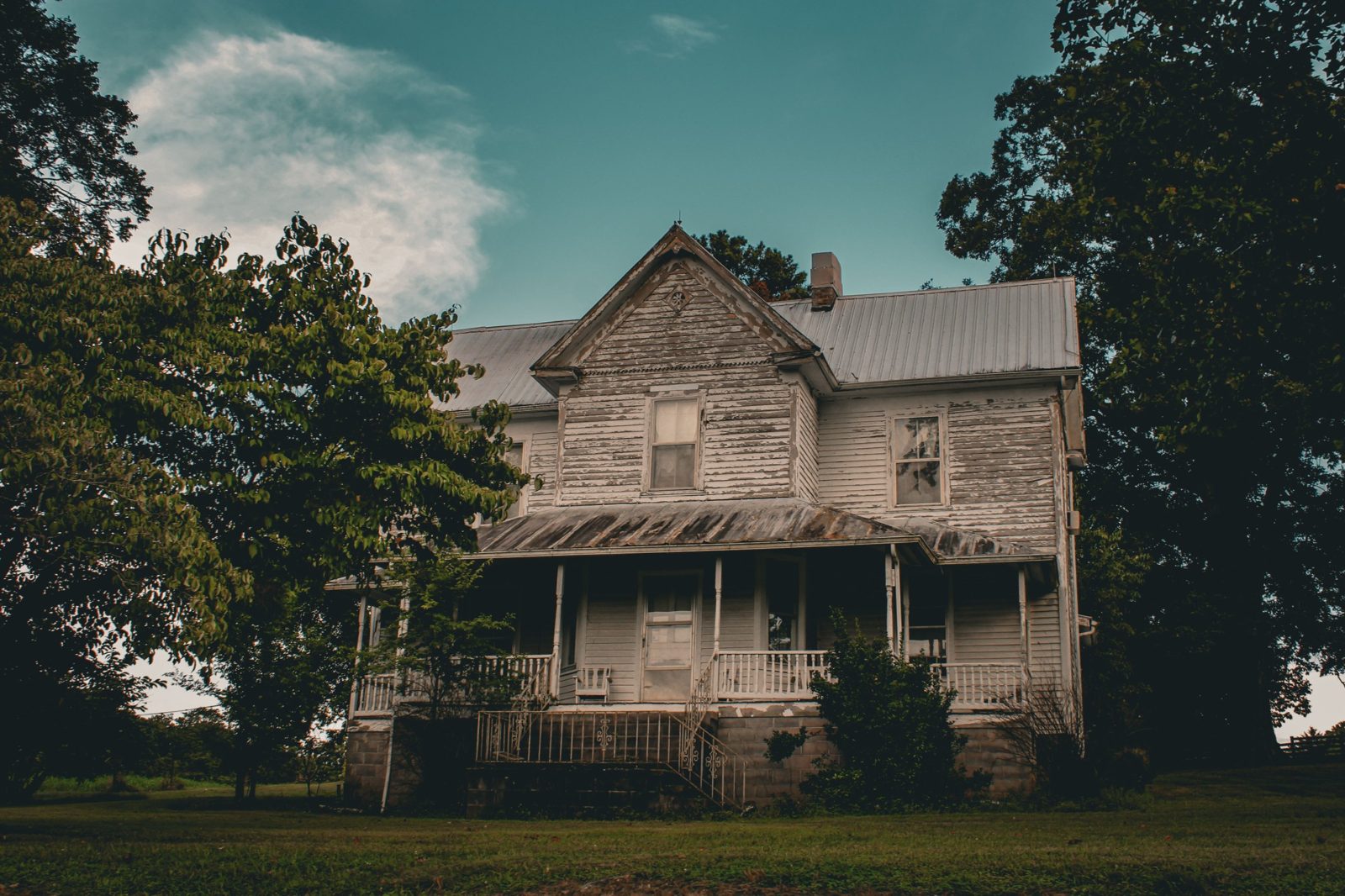 Weathered two-story farmhouse with peeling white paint and a wraparound porch under a teal sky